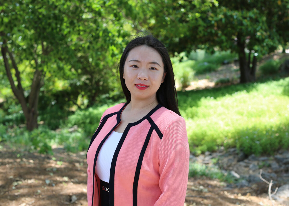 Fan Zhang in a white shirt an dpink blazer with black trim, outdoors in front of green shrubs.