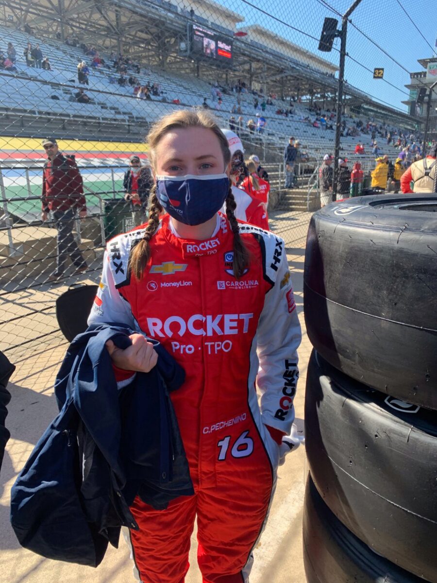 Chelsea in a team firesuit next to a stack of tires in the pit. Her hair is in braids.